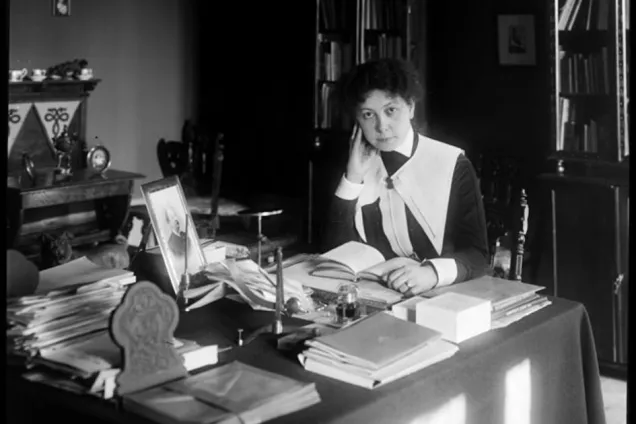 Svartvitt foto. Valfrid Palmgren sitter vid ett skrivbord, fullt av papper och böcker i högar.